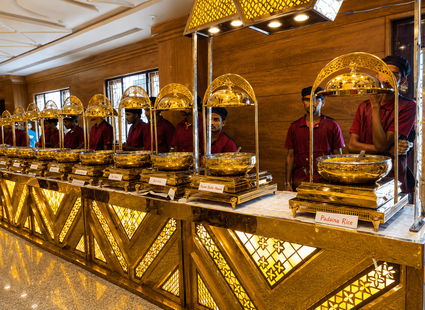 Staff in maroon uniforms serving from an elegant gold buffet counter with domed chafing dishes at a Jayram Catering live counter setup