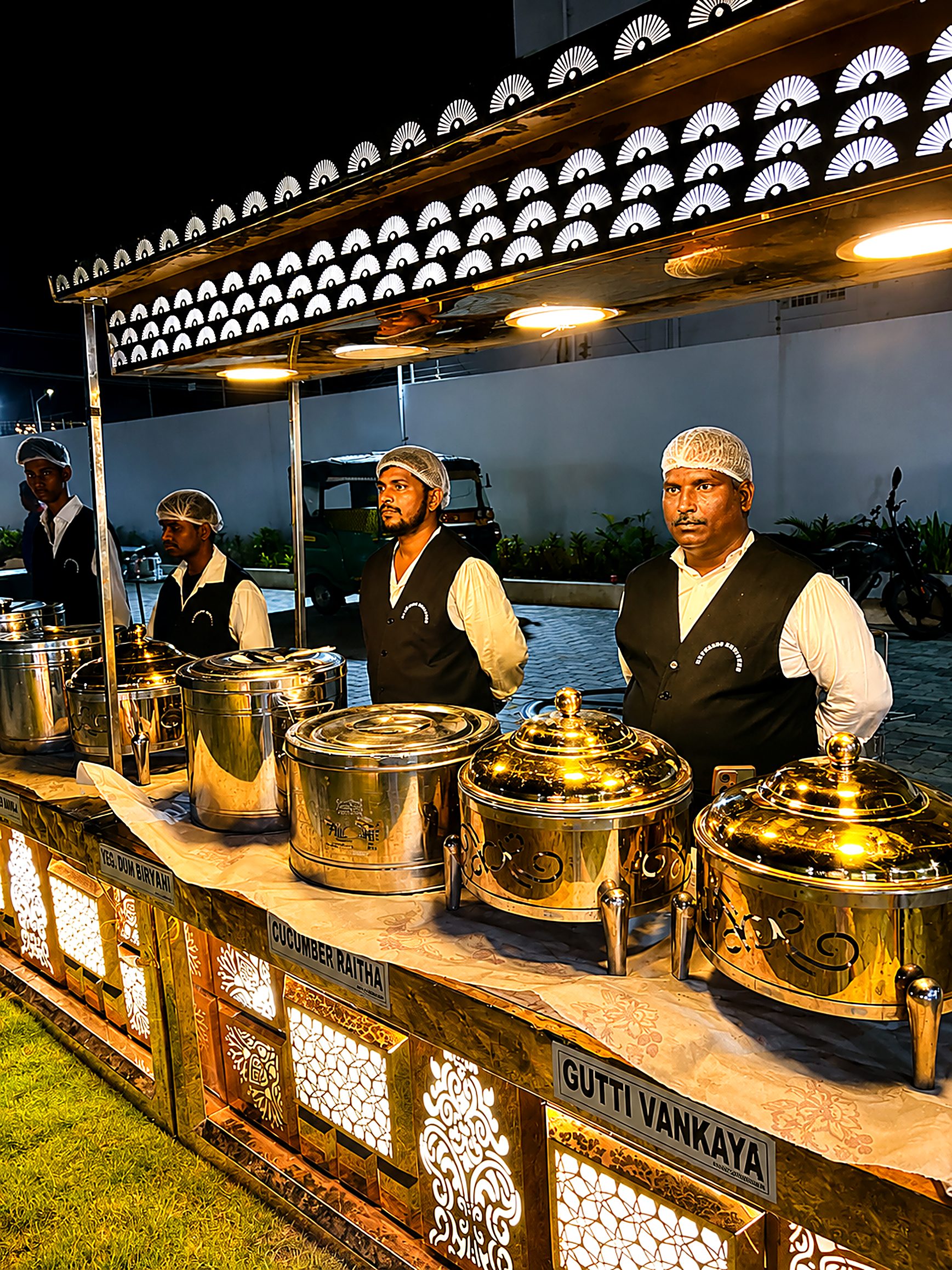 Uniformed live-counter staff serving from gleaming gold chafing dishes at an evening Jayram Catering event
